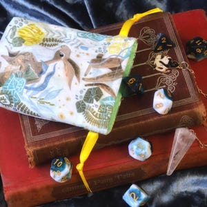 May include: A fabric drawstring pouch with a light-coloured print of rabbits and foliage, featuring a bright yellow ribbon closure. The pouch rests on two stacked hardcover books alongside several multi-sided gaming dice and a clear crystal pendulum.