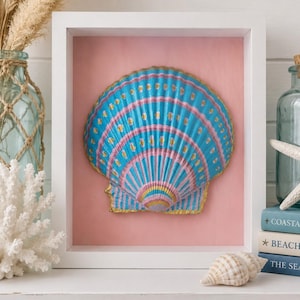 May include: A white shadow box frame containing a single large scallop shell painted with blue, pink, and gold stripes and dots. The shell is centered against a solid light pink background, creating a decorative wall art piece for display.