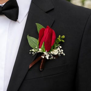 May include: A boutonniere featuring a single deep red rosebud accented with small white baby's breath flowers and green leaves. The stem is wrapped in brown ribbon, pinned to the lapel of a black suit jacket worn with a white shirt and black bow tie.