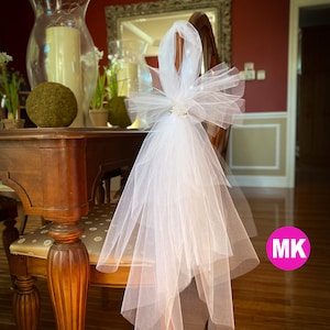 May include: White tulle chair sash with a large bow and a rhinestone embellishment. The sash is tied around the back of a wooden chair.