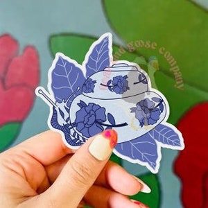 May include: A blue and white teacup snail sticker with floral details. The sticker is on a hand against a background of red and green flowers.