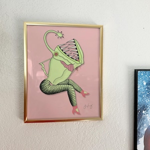 May include: A framed illustration of a green anglerfish with a human-like body wearing fishnet stockings and high heels on a pink background. The anglerfish has a large mouth with sharp teeth and a glowing lure on its head. The illustration is signed "J+G".