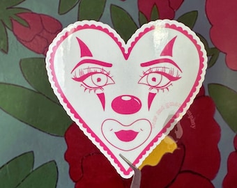 May include: Pink heart-shaped sticker with a clown face. The clown has large eyes, a red nose, and a pink smile. The sticker has a scalloped edge and is on a white background.