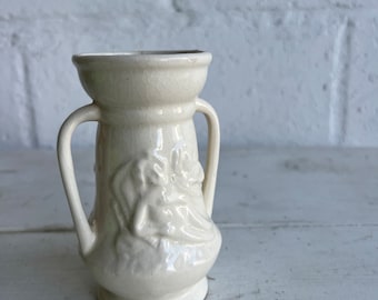 May include: A small, white ceramic vase with two handles and a floral design. The vase has a textured surface and a glossy finish.