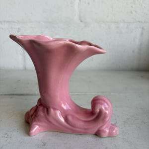 May include: A pink ceramic cornucopia vase featuring a flared, wavy rim and a curved, horn-shaped body. The base of the vessel is decorated with molded, leaf-like scrollwork details that extend from the bottom of the horn to the curled tip.