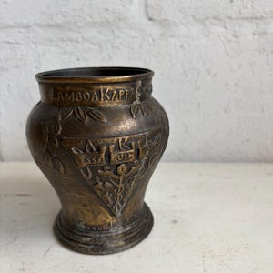 May include: A bronze-colored, antique-style vase with embossed details. The vase features the text "LAMBDA KAPPA" around the rim and a triangular emblem with additional text and symbols. The vase has a flared base and a slightly wider body.