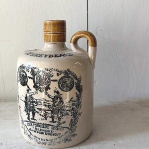 May include: Cream-colored stoneware jug with a brown handle and stopper. The jug features a black and white illustration of two figures in traditional Scottish attire, with text that reads "Heather Dew Blended Scotch Whisky."