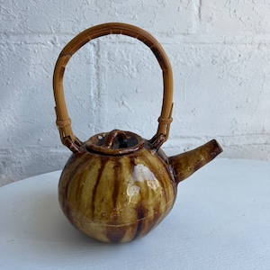 May include: A ceramic teapot with a rounded body and a bamboo handle. The teapot is glazed in shades of brown with vertical streaks. It has a spout and a small lid. The teapot is sitting on a white surface.