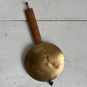 May include: A vintage clock pendulum with a round, brass disc and a wooden rod. The brass disc has a slightly tarnished appearance, and the wooden rod is a warm brown color. The pendulum is designed for a clock mechanism.
