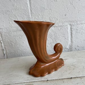 May include: A brown ceramic vase shaped like a horn with a fluted design. The vase has a wide opening and a curled handle.