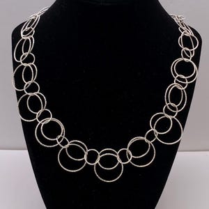 Puede incluir: Collar plateado con una serie de círculos abiertos interconectados. Los círculos varían en tamaño y tienen un acabado texturizado. El collar se exhibe en un maniquí negro, resaltando su diseño y brillo.