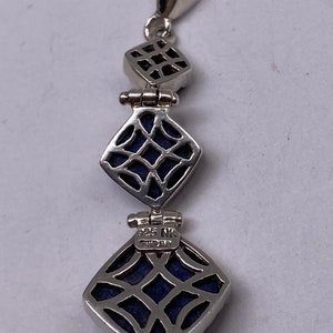 May include: A silver pendant with three square-shaped lapis lazuli stones. The stones are set in a silver frame with a delicate filigree design. The pendant has a silver bail and is marked "925 NK" on the back.
