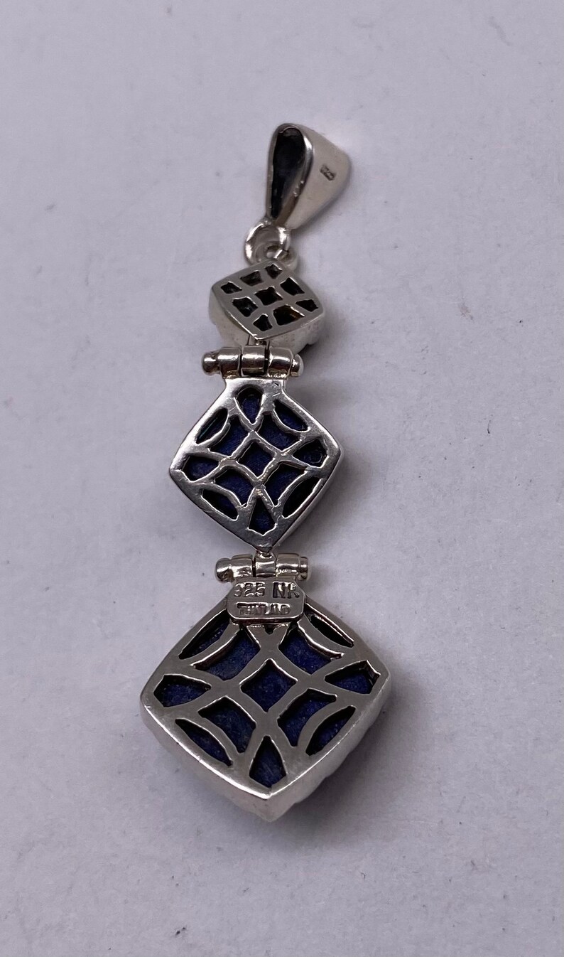 May include: A silver pendant with three square-shaped lapis lazuli stones. The stones are set in a silver frame with a delicate filigree design. The pendant has a silver bail and is marked "925 NK" on the back.