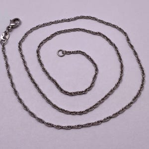 Op de afbeelding: Een zilverkleurige touwketting met een karabijnsluiting. De ketting is in een spiraalvorm gewikkeld, wat het ingewikkelde gedraaide ontwerp laat zien. De ketting is gemaakt van een glanzend, metalen materiaal.