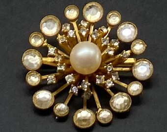 May include: A gold tone brooch with a large pearl in the center and many smaller rhinestones radiating out from the pearl.