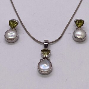May include: A silver necklace and earring set featuring a pendant and two earrings. Each piece has a round, white pearl and a triangular, light green gemstone. The jewelry is set against a white background.