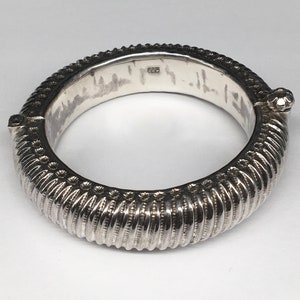 May include: A silver bangle bracelet with a textured design. The bracelet is hinged and has a small decorative knob on the side.