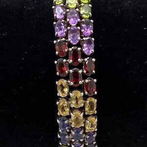 May include: A multi-gemstone bracelet featuring rows of oval-cut gemstones in various colors, including purple, green, yellow, and red. The bracelet is set in a silver-toned metal and is displayed against a black background.