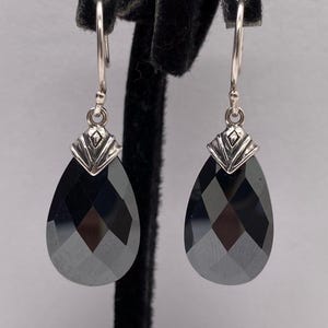 May include: A pair of silver-tone earrings with a teardrop-shaped, faceted, black gemstone. The earrings hang from a silver hook and have a decorative silver top. The earrings are displayed on a black velvet stand.
