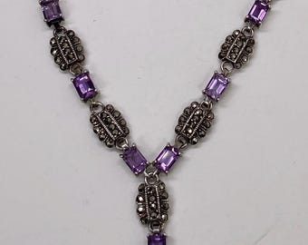 Può includere: Una collana argentata con pietre preziose rettangolari viola e elementi metallici ovali decorativi. Il design a forma di Y presenta un pendente centrale. Le pietre sono di un viola intenso, su sfondo bianco.