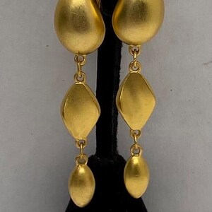 Puede incluir: Pendientes colgantes dorados con forma de lágrima en la parte superior, seguidos de un centro en forma de diamante y una parte inferior ovalada. Los pendientes cuelgan de un expositor negro. La joyería es probablemente de metal.