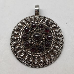 May include: A silver pendant with a circular design featuring a central cluster of 12 dark red gemstones surrounded by a decorative border.