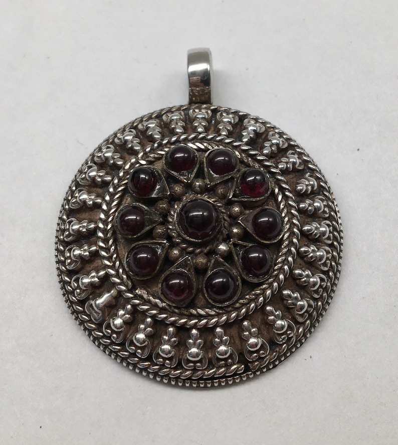 May include: A silver pendant with a circular design featuring a central cluster of 12 dark red gemstones surrounded by a decorative border.