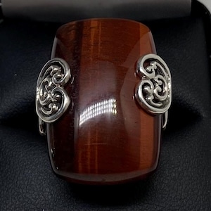 May include: A cylindrical, reddish-brown bracelet with a polished finish. The bracelet features ornate, silver-colored metal accents on each side. The design is symmetrical and the overall aesthetic is elegant and refined.