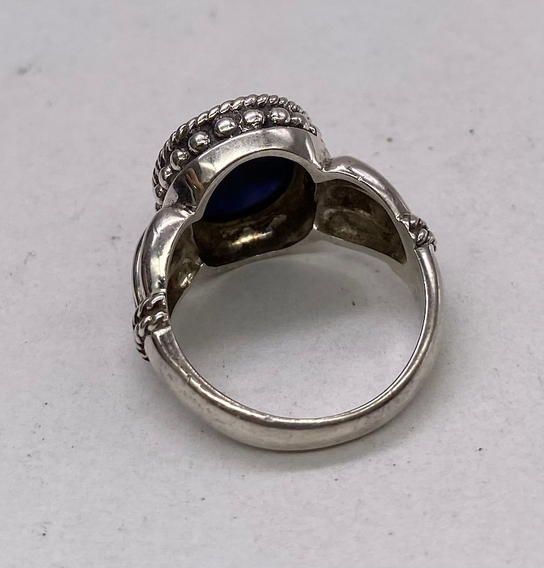 Peut inclure: Une bague en argent avec une pierre pr&eacute;cieuse bleue sertie dans un sertissage &agrave; b&eacute;li&egrave;re. La bague a une bande d&eacute;corative avec un motif en forme de corde.