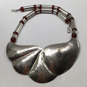 May include: A silver necklace with a large, hammered, leaf-shaped pendant. The necklace is made of silver beads and red carnelian beads.