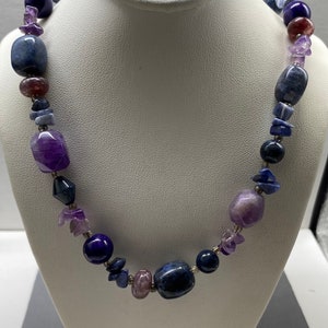 May include: A necklace made of various shades of purple and blue gemstones. The necklace features a variety of shapes and sizes of stones, including round, oval, and rectangular.