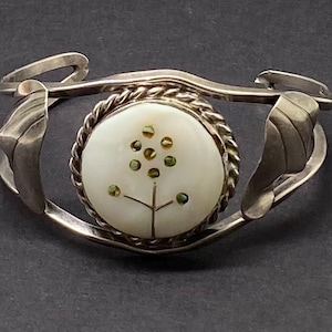 May include: A silver bracelet with a round, white mother-of-pearl inlay featuring a stylized tree with green accents.