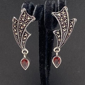 May include: A pair of silver earrings with a dangling red gemstone. The earrings are made of a delicate, intricate design with a floral motif.