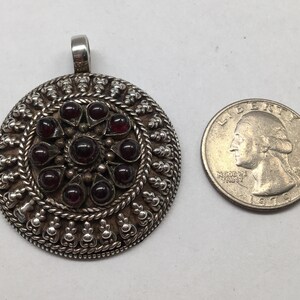 May include: A silver pendant with a circular design featuring a central cluster of red gemstones surrounded by intricate silver filigree. The pendant is approximately the size of a US quarter.