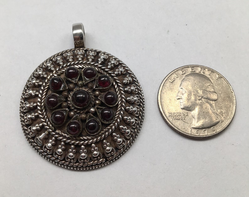 May include: A silver pendant with a circular design featuring a central cluster of red gemstones surrounded by intricate silver filigree. The pendant is approximately the size of a US quarter.