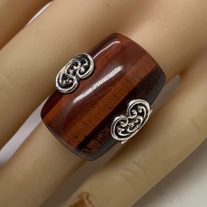 May include: A wide, rectangular ring with a polished, reddish-brown stone. The ring features two ornate, silver-colored embellishments on the sides. The stone has a striped pattern, and the ring is set on a light-colored background.