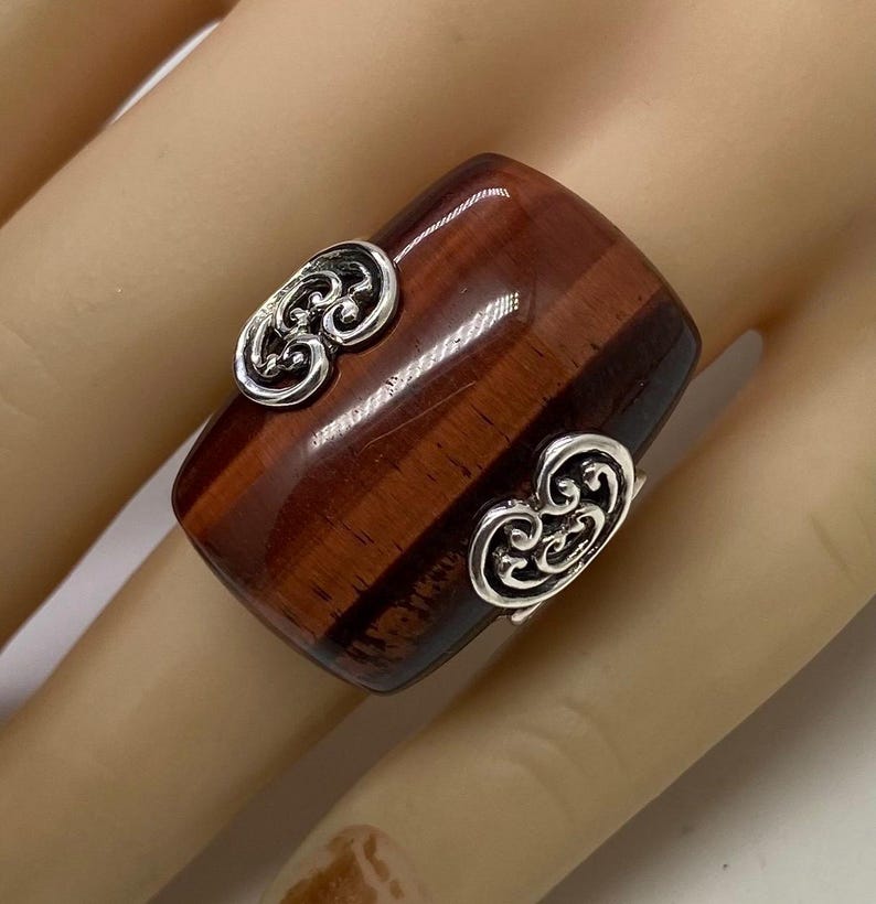 May include: A wide, rectangular ring with a polished, reddish-brown stone. The ring features two ornate, silver-colored embellishments on the sides. The stone has a striped pattern, and the ring is set on a light-colored background.