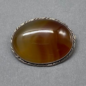 May include: An oval-shaped agate brooch with a brown, orange, and yellow banded pattern. The brooch is set in a silver-toned metal with a rope-like border.