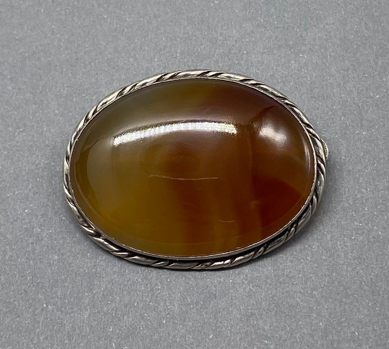 May include: An oval-shaped agate brooch with a brown, orange, and yellow banded pattern. The brooch is set in a silver-toned metal with a rope-like border.