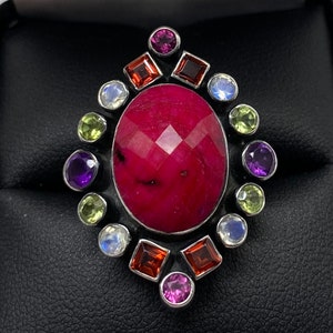 May include: A silver ring with a large, faceted, pink gemstone surrounded by smaller gemstones in various colors, including green, purple, red, and white.