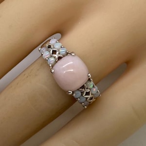 K&ouml;nnte beinhalten: Ein silberner Ring mit einem gro&szlig;en rosafarbenen Opal-Edelstein, umgeben von kleineren wei&szlig;en Opalen.