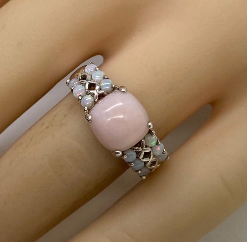 K&ouml;nnte beinhalten: Ein silberner Ring mit einem gro&szlig;en rosafarbenen Opal-Edelstein, umgeben von kleineren wei&szlig;en Opalen.