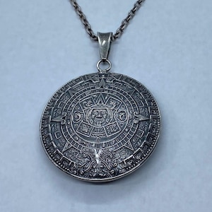 May include: A silver-toned pendant necklace featuring a detailed Aztec calendar design. The circular pendant showcases intricate carvings of figures and symbols, suspended from a delicate chain. The pendant is approximately 2 inches in diameter.