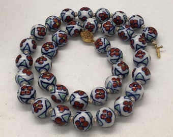 May include: A white ceramic necklace with red and blue floral designs on each bead. The necklace is strung with a gold clasp.