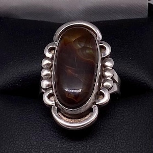 May include: A silver ring featuring an oval, brown gemstone with dark streaks. The ring has a decorative silver setting with curved and beaded accents, showcasing a vintage or artisan design.