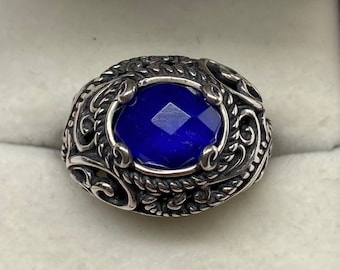 Könnte beinhalten: Ein silberner Ring mit einem verzierten Design, das einen großen, facettierten, blauen Edelstein zeigt.