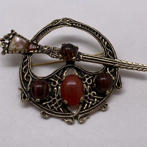May include: A vintage brooch with a gold-tone metal filigree design. The brooch features a central oval red stone, surrounded by three brown, round stones. A decorative pin with a patterned head and a decorative shaft is set across the brooch.