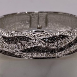 May include: Silver-toned bangle watch with a wave-like pattern of clear and black crystals. The watch face is silver with a small crown. The bracelet is set on a white surface.