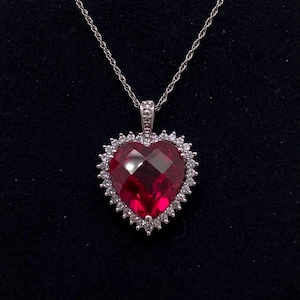 May include: A silver necklace with a heart-shaped pendant. The pendant has a large, faceted red gemstone surrounded by a halo of smaller, clear gemstones. The chain is delicate and silver in colour.