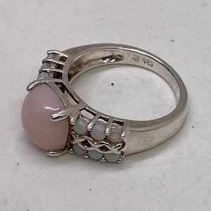 K&ouml;nnte beinhalten: Ein silberner Ring mit einem gro&szlig;en rosafarbenen Opal-Edelstein und kleineren wei&szlig;en Opalen, die in einem filigranen Design eingefasst sind.
